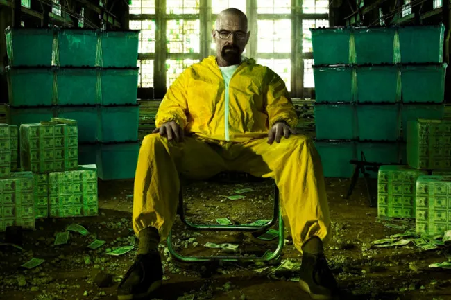 Walter White from Breaking Bad, in a yellow hazmat suit, sits surrounded by stacks of money and green containers.
