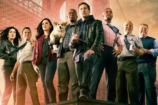 The main cast of the TV show Brooklyn Nine-Nine posing, with Cheddar the corgi included, under a bridge.