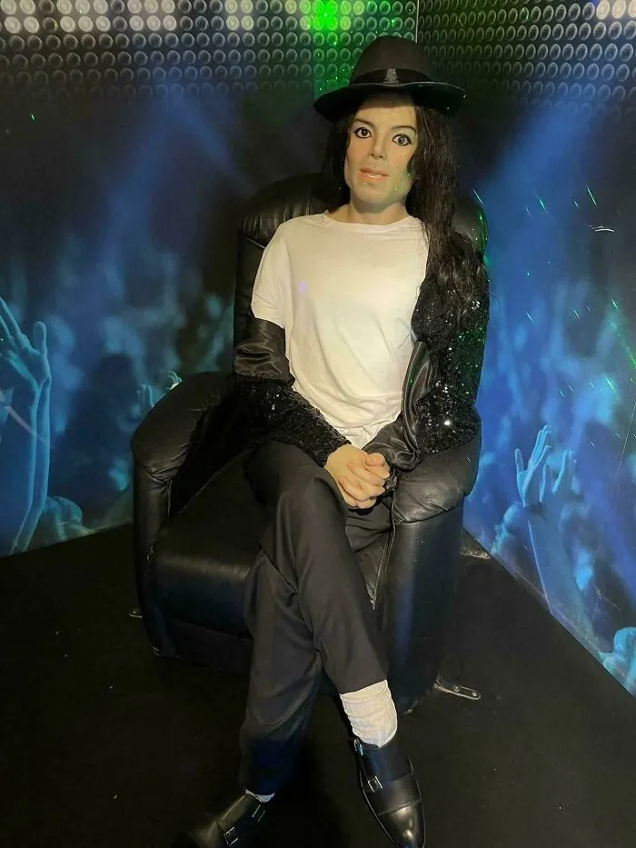 Wax figure of a celebrity wearing a fedora and black and white outfit seated in chair