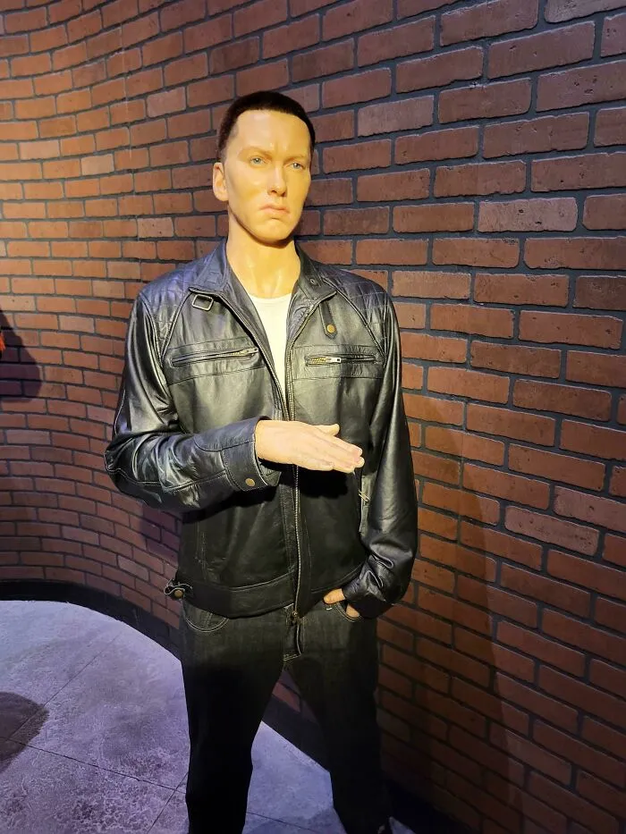 Celebrity wax figure wearing black leather jacket with hand gesture near brick wall