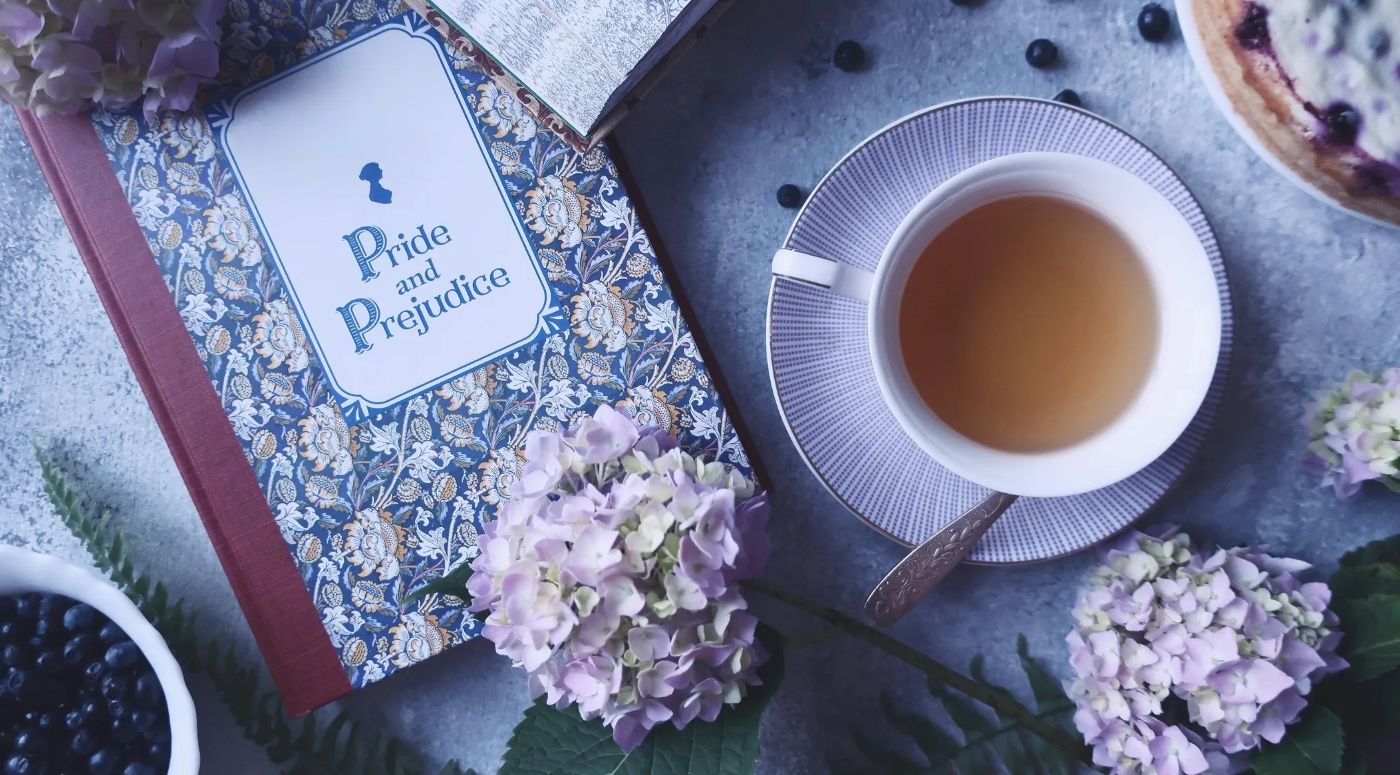 A copy of Pride and Prejudice next to a cup of tea, with hydrangeas and blueberries on a gray surface. General Knowledge Quiz.