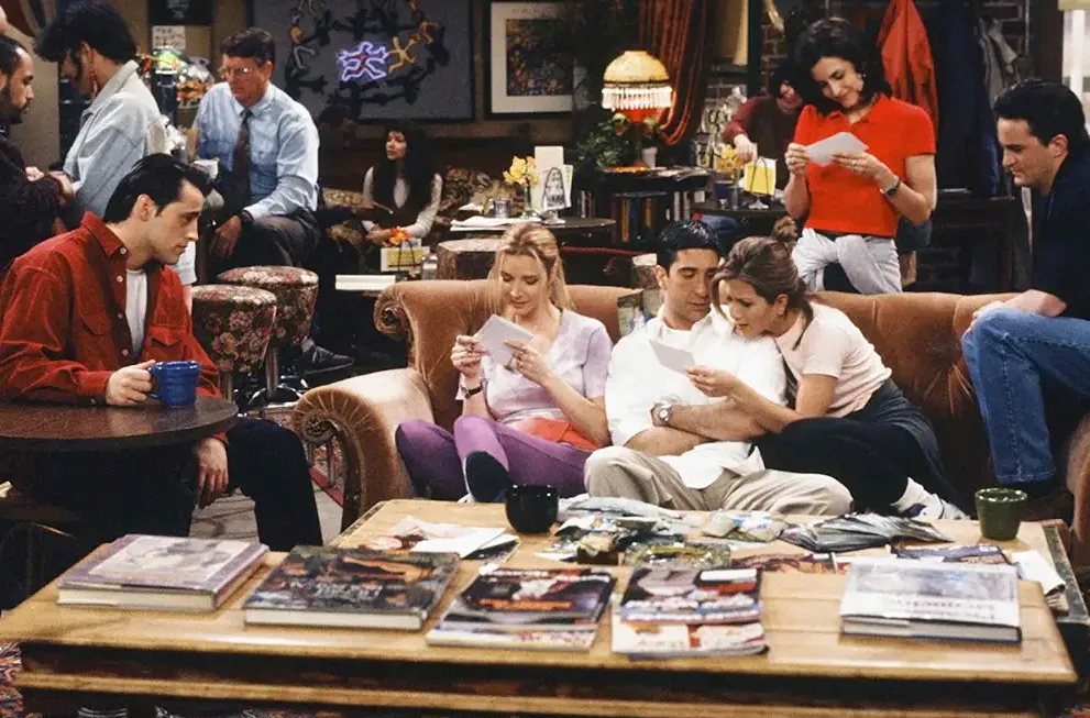 Friends characters in the iconic coffee shop. Monica, Phoebe, Rachel, Ross, Joey, and Chandler are reading papers, playing a general knowledge quiz.