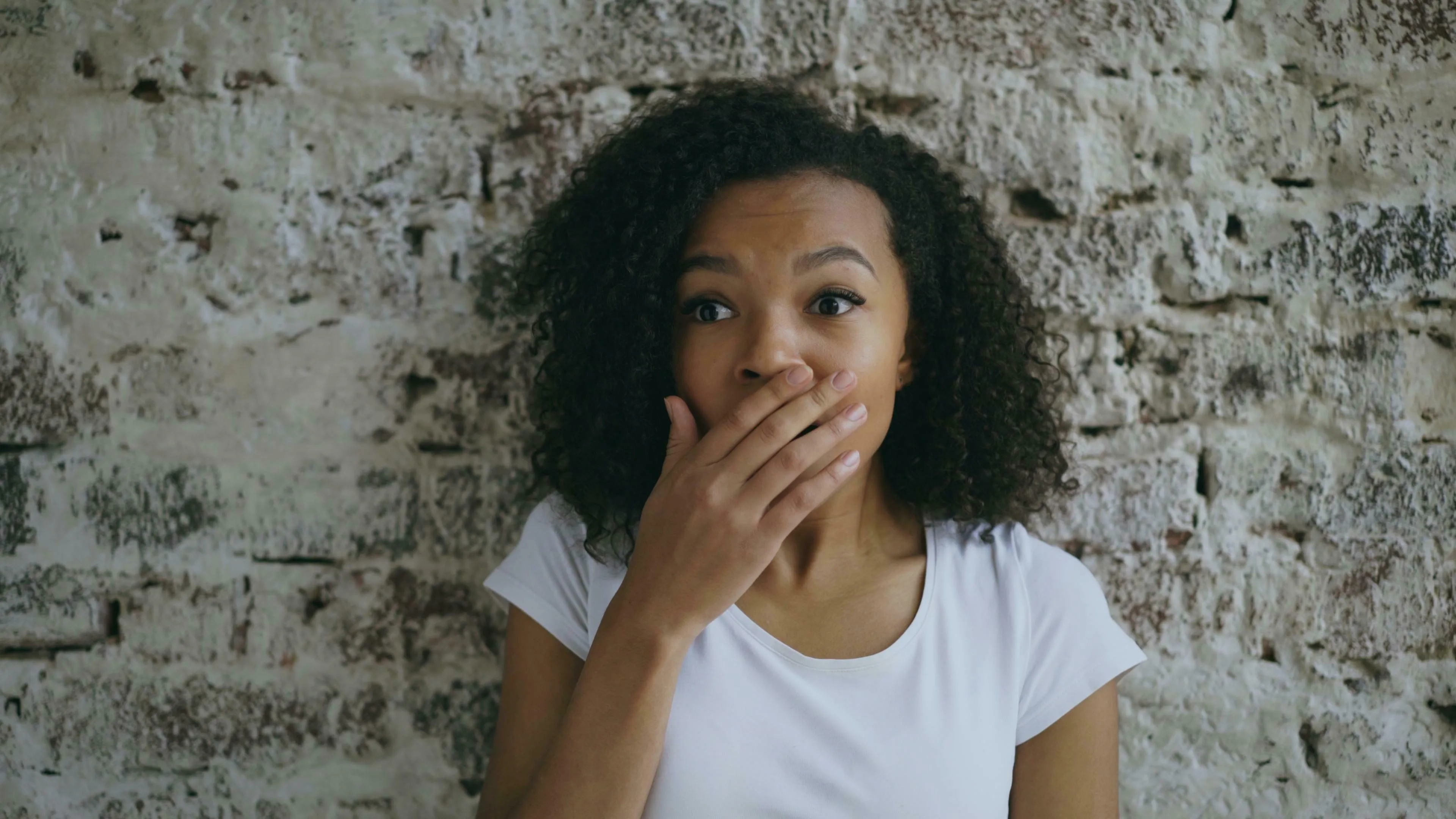 A woman with curly hair covers her mouth, looking surprised or worried against a stone wall. How harsh are you on yourself after a mistake?