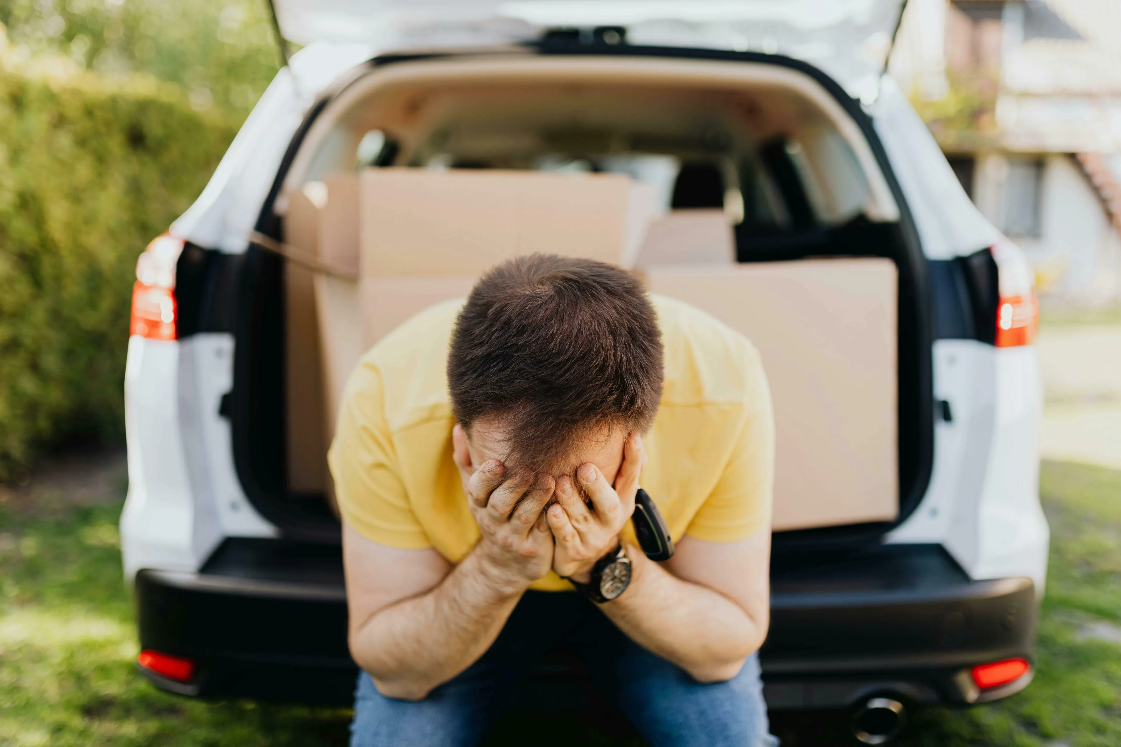 A man with his face in his hands in front of an open car trunk with boxes, depicting a bad day.