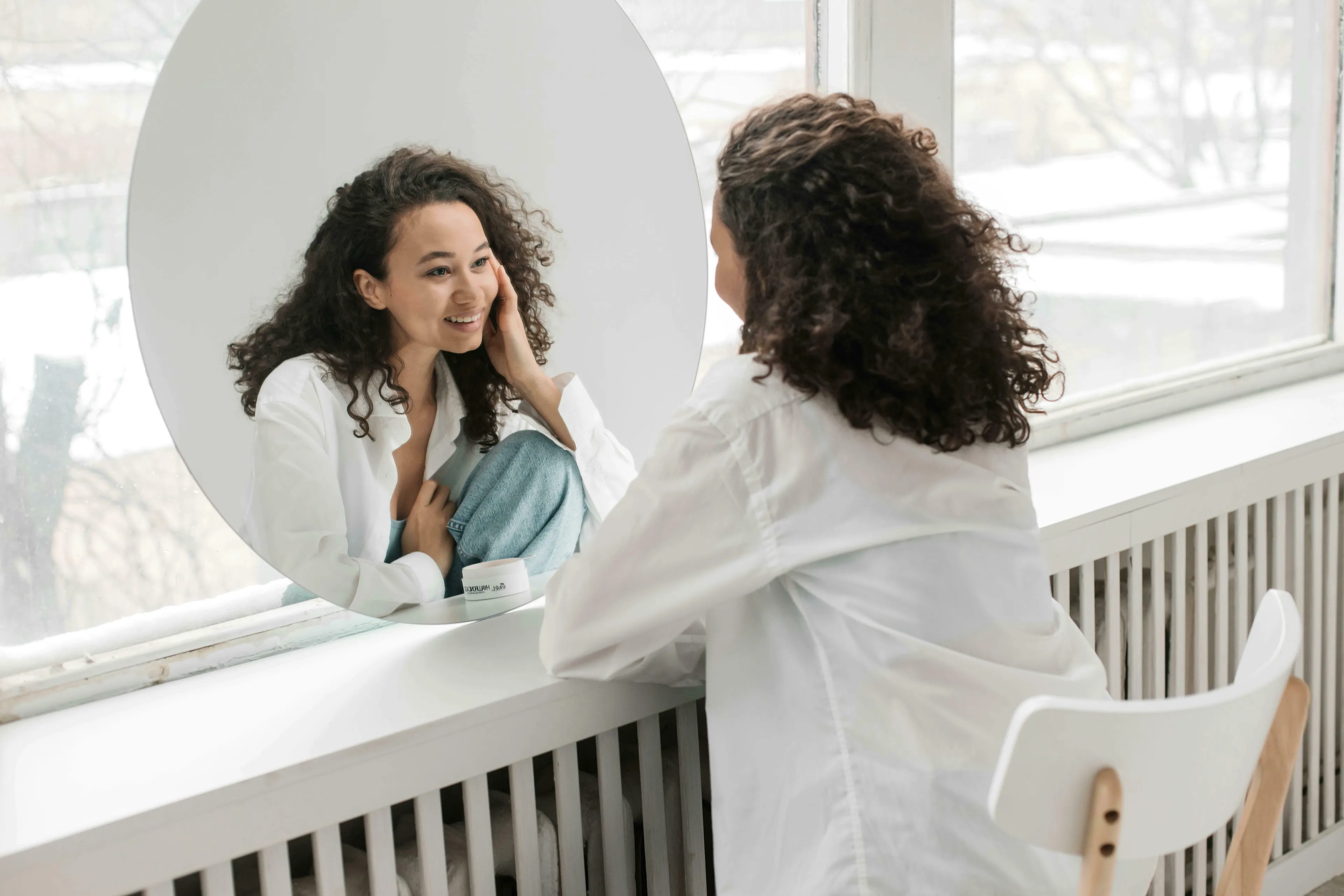 Woman with curly hair looking in a mirror and smiling, contemplating how harsh she is on herself and self-treatment.