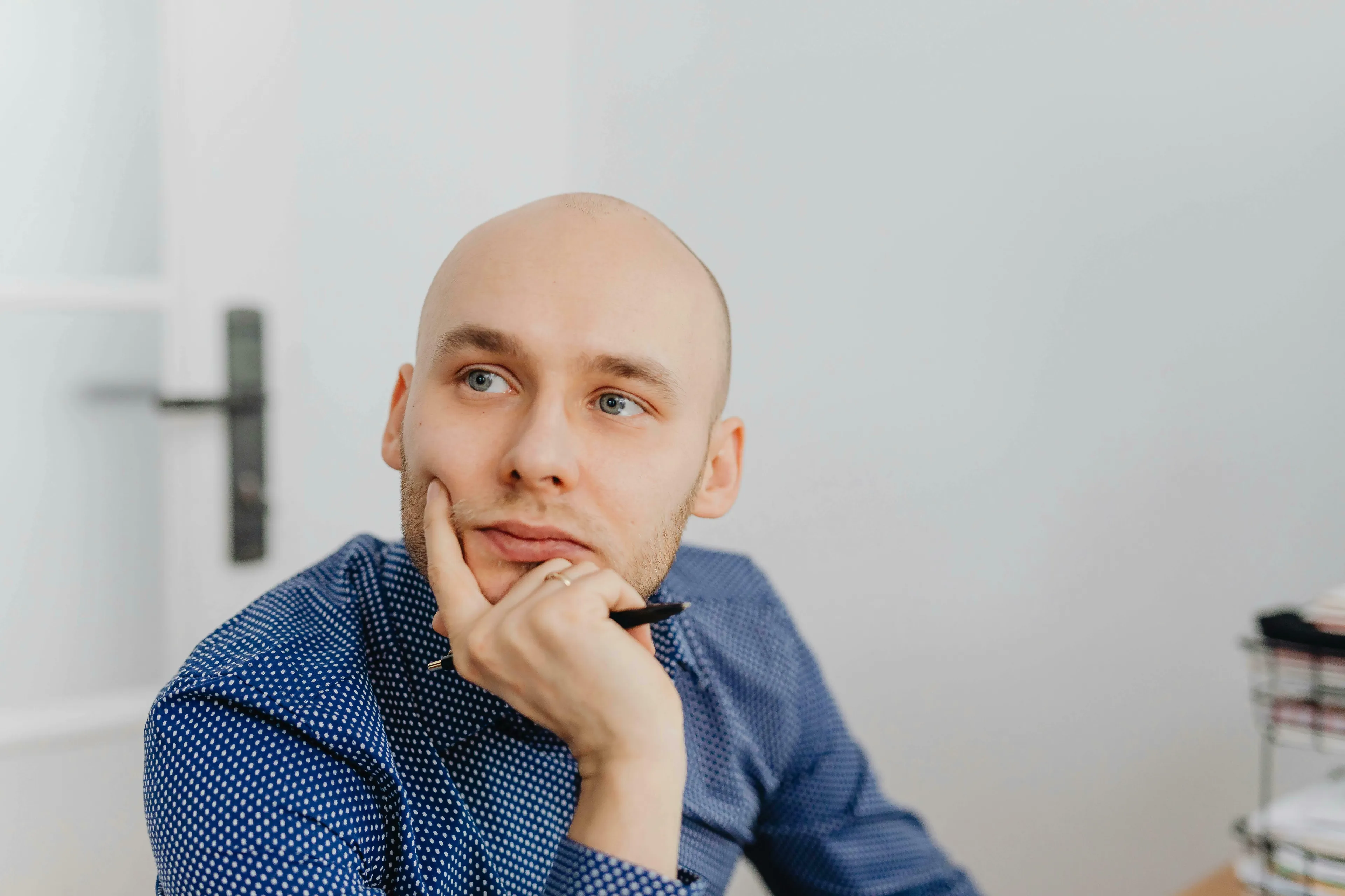 A pensive bald man in a blue polka-dot shirt, holding a pen, reflecting on how harsh are you on yourself.