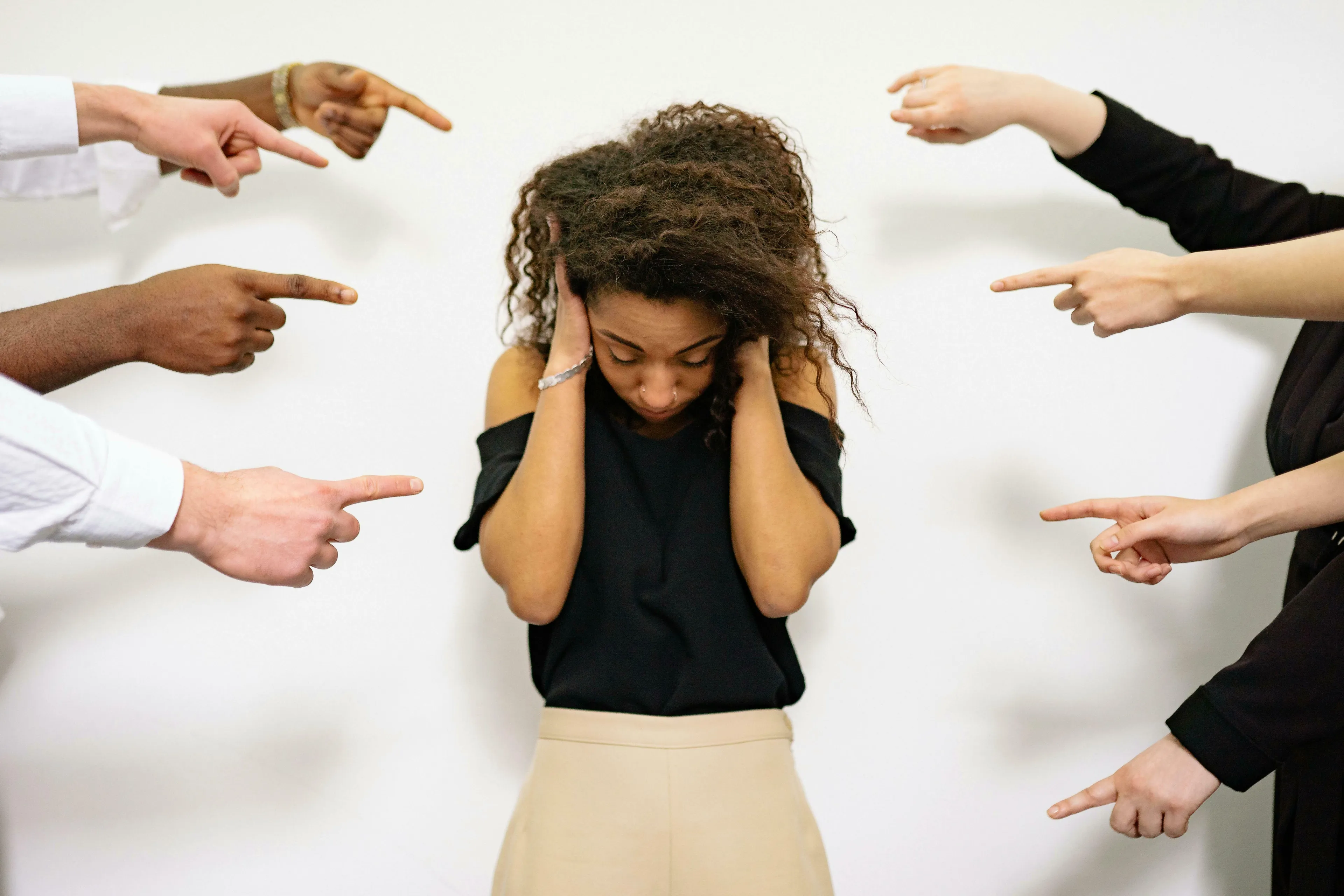 A young woman with curly hair covers her ears, looking down, as multiple hands point accusatorily at her, depicting how harsh criticism feels.