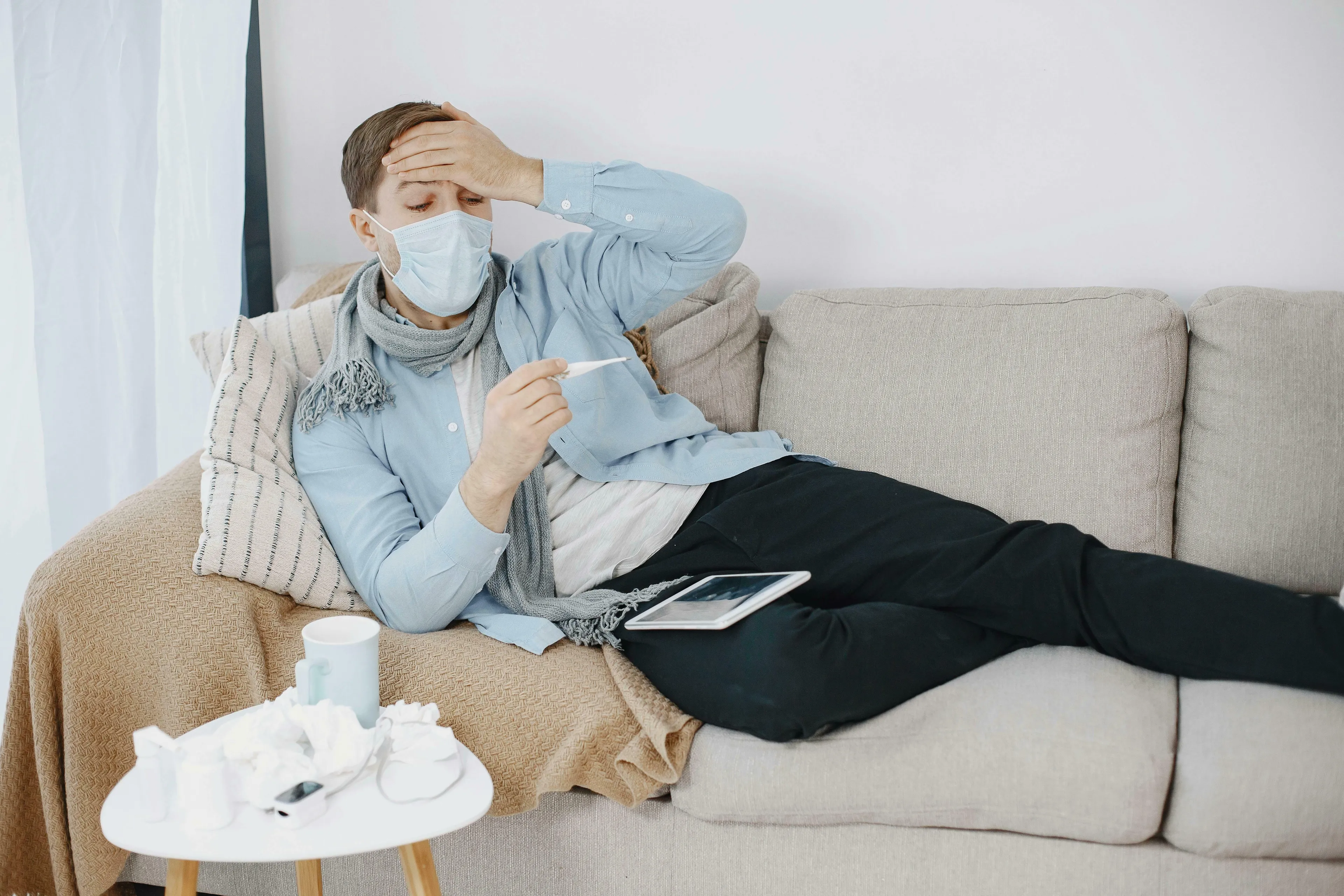 A man in a mask and scarf, feeling unwell on a couch, checking his temperature. He looks sick. How harsh are you on yourself?