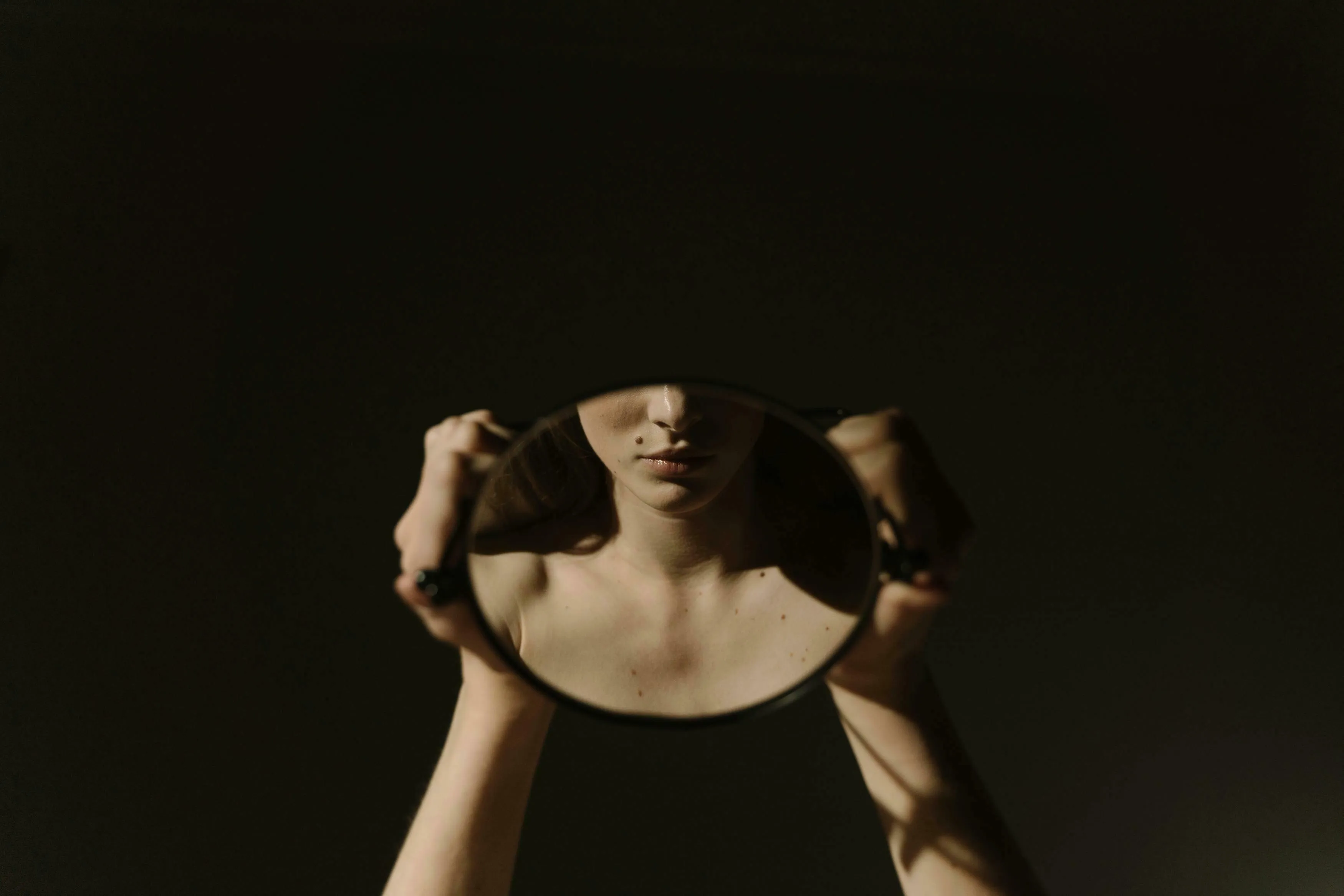A person holds a mirror reflecting their chest and lips, with dark lighting. How harsh are you on yourself?