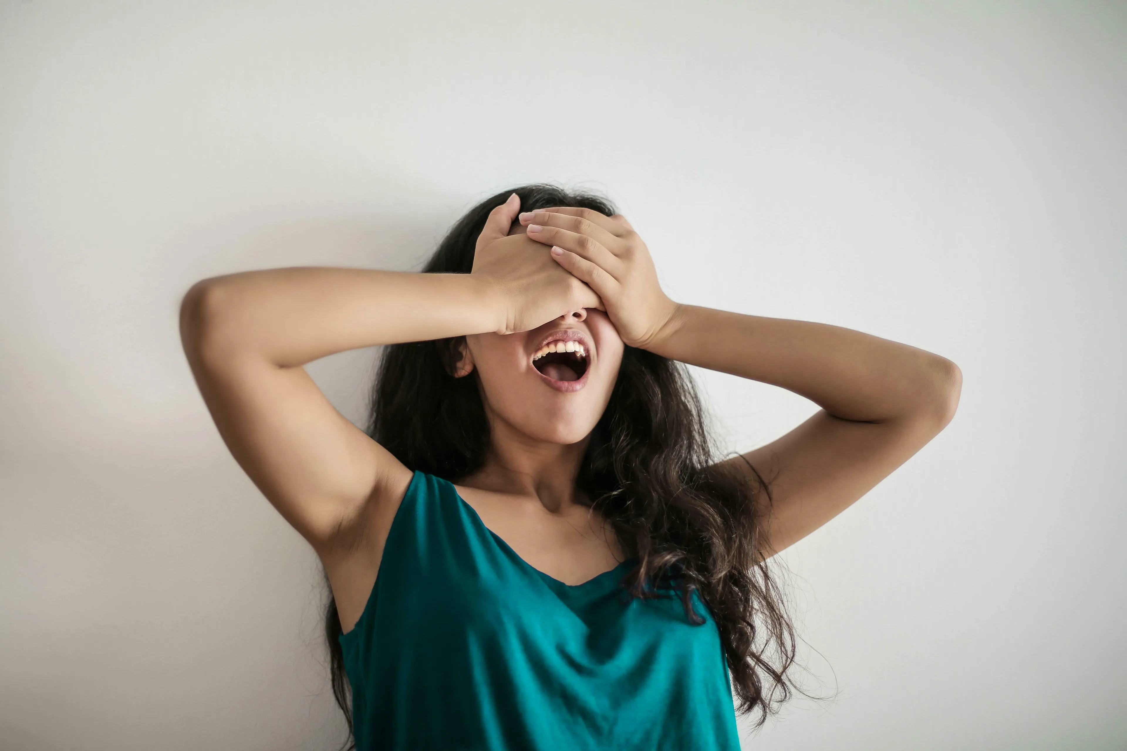 A woman in a teal top covers her eyes with hands, mouth open, against a white wall. How harsh are you on yourself?