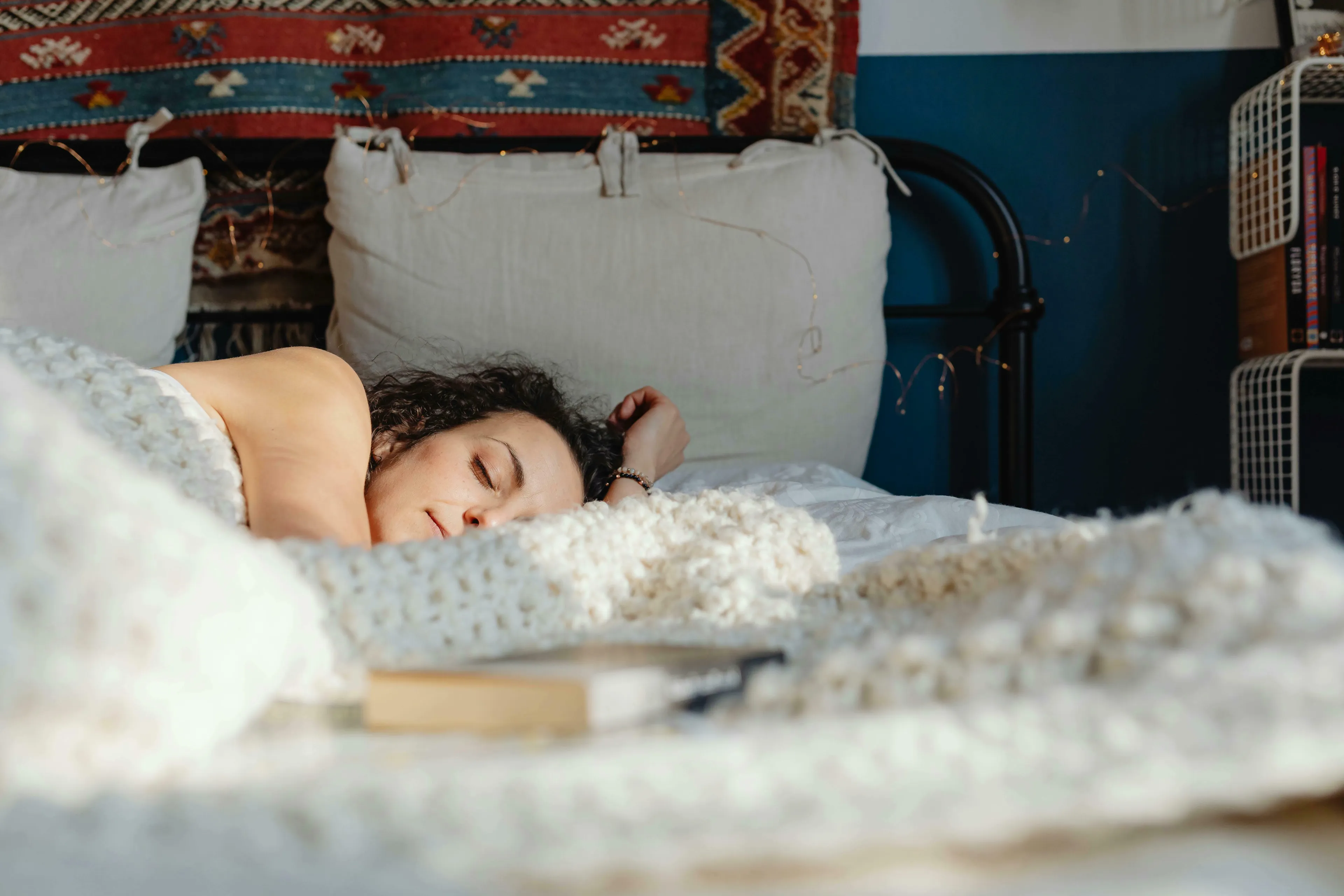 A woman with curly hair peacefully sleeping under a warm, chunky knit blanket, showing what rest feels like. How harsh are you on yourself?