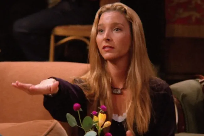 Phoebe Buffay, a fictional character with long blonde hair, gesturing with an open palm. She wears a dark top and necklace.