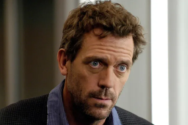 A close-up of Dr. House looking intensely forward, with blue eyes and light brown messy hair. He wears a striped shirt.