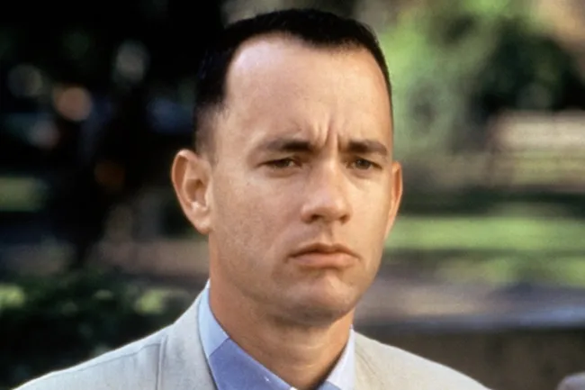 Close-up of Forrest Gump, portrayed by Tom Hanks, looking serious with a light blue shirt and beige jacket.