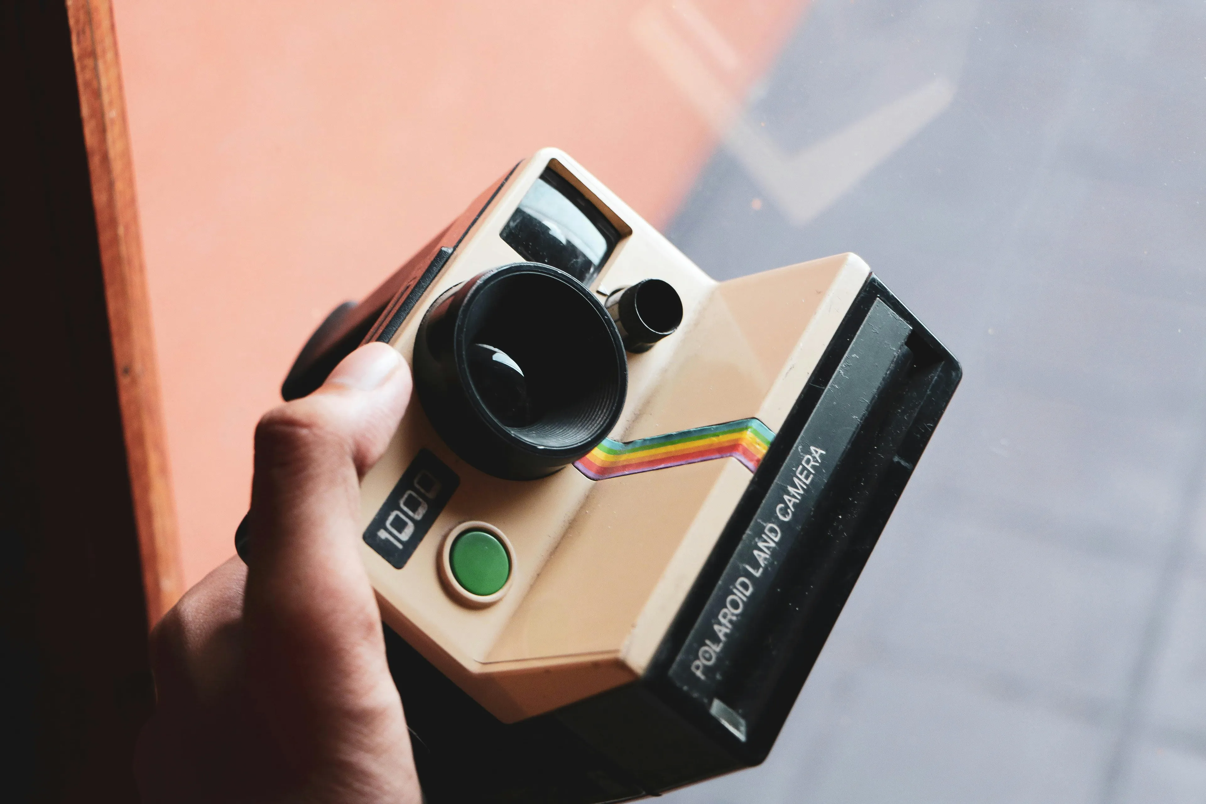 A person holding a vintage Polaroid camera, featuring its classic beige body, green button, and rainbow stripe.