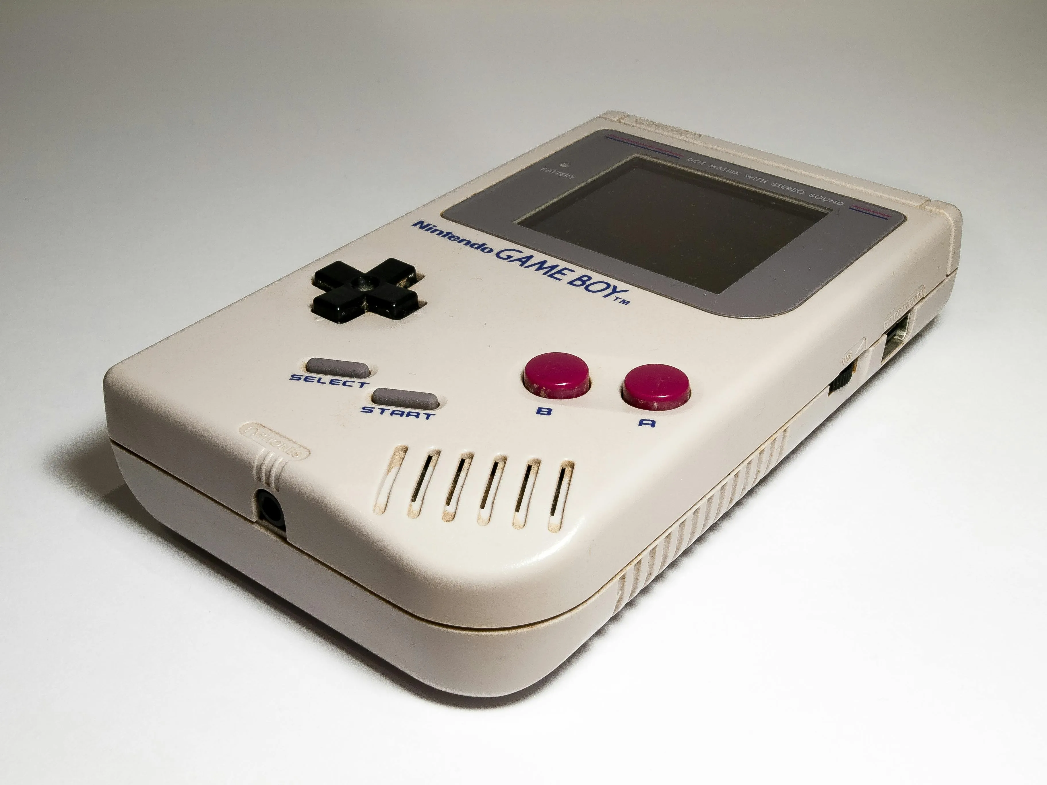 An old, grey Game Boy on a white surface. It features black directional pad and red A and B buttons.