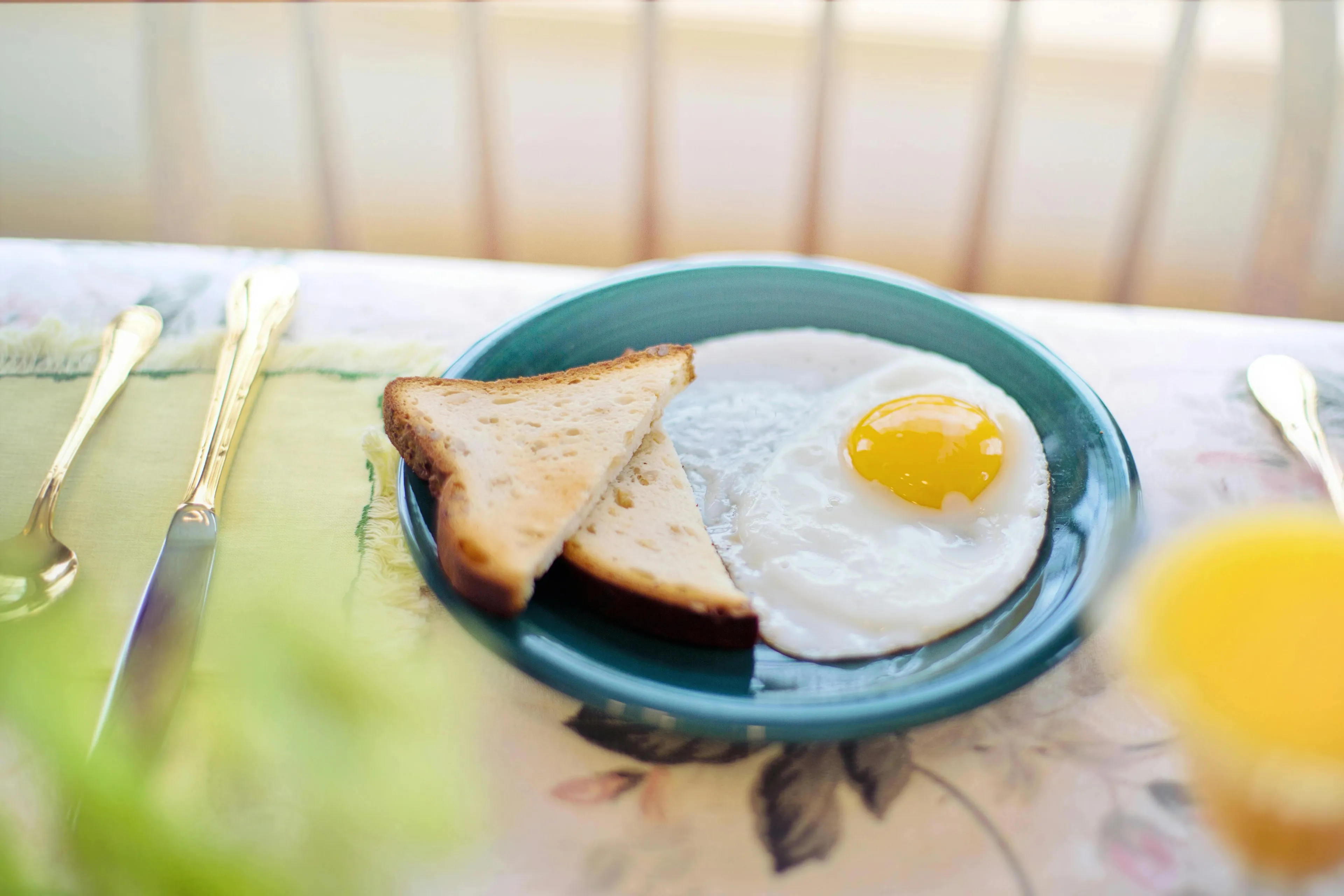 A blue plate with a fried egg and two slices of toast on a floral tablecloth, with golden cutlery. Eggs and toast for morning energy.