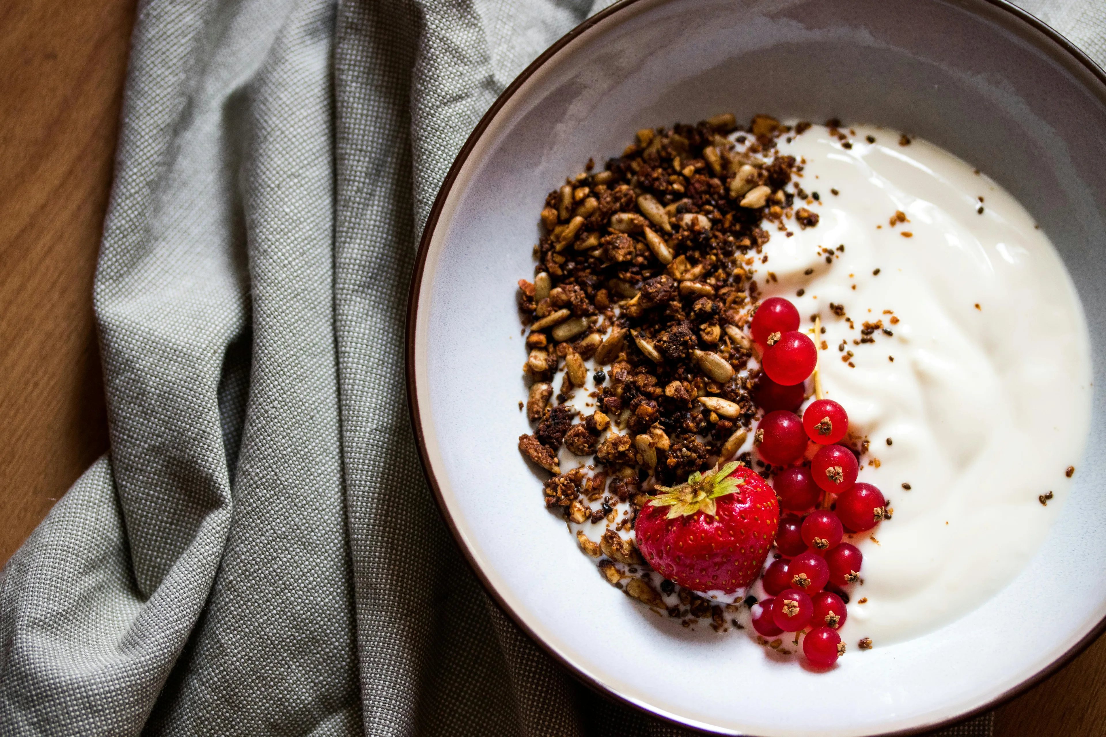A bowl of granola with yogurt, topped with red currants and a fresh strawberry, on a textured grey cloth.