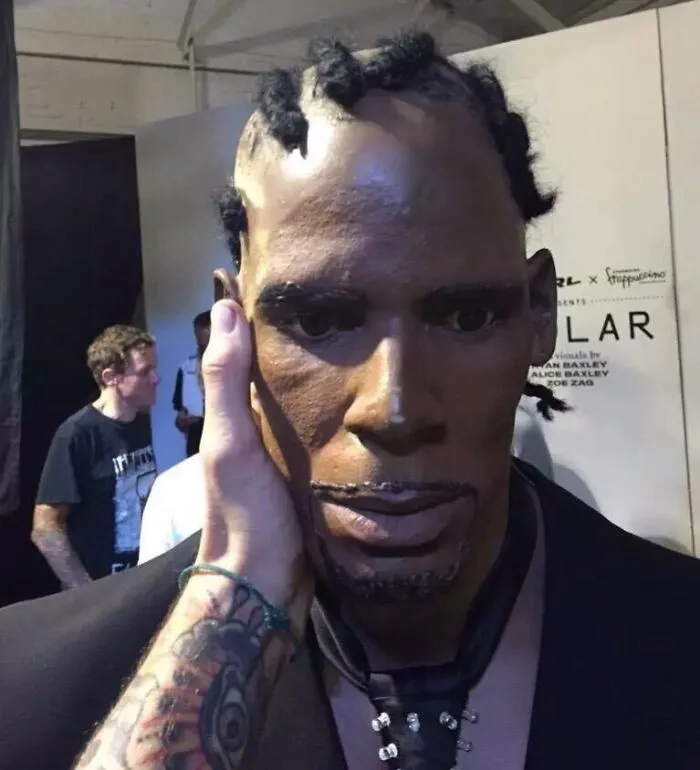 Uncanny celebrity wax figure with detailed facial features and braided hair held by a tattooed hand