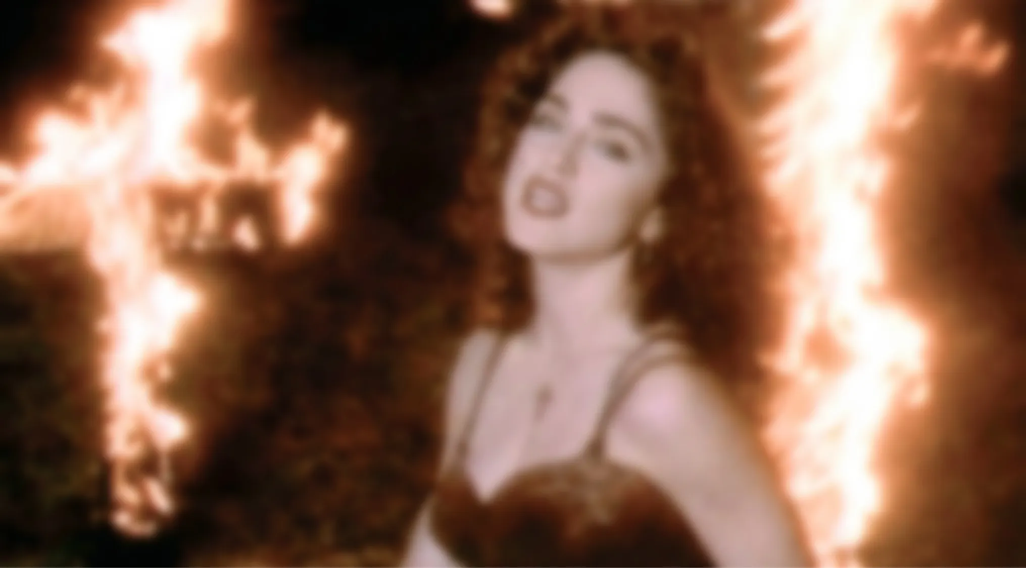 A blurry music video image of a woman with curly hair and a dark top, with flames in the background. Blurry Music Videos Quiz.