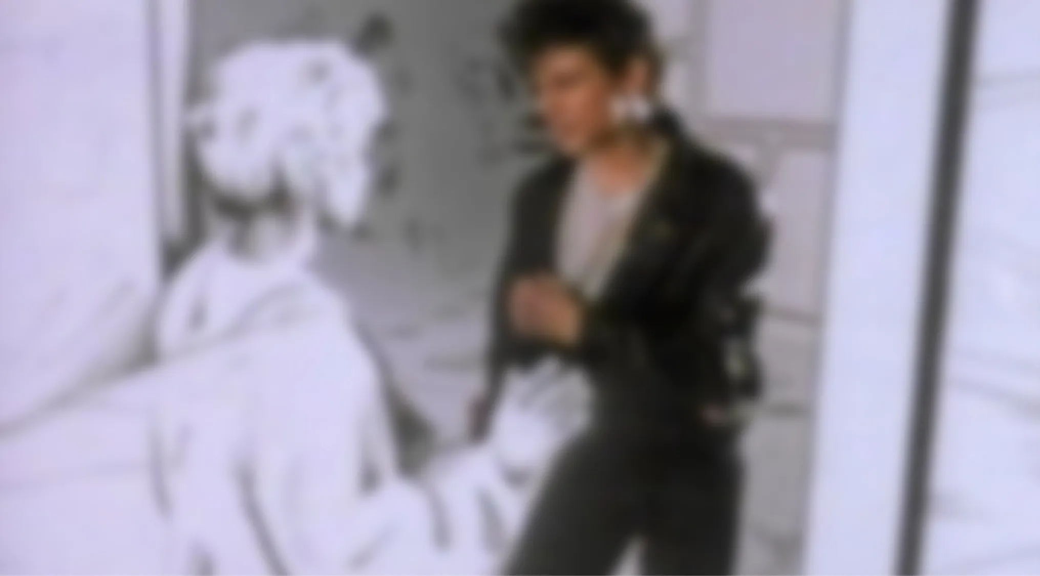 A blurry music video image of a person in a dark jacket and light shirt standing, facing another figure. Which music video is shown here?