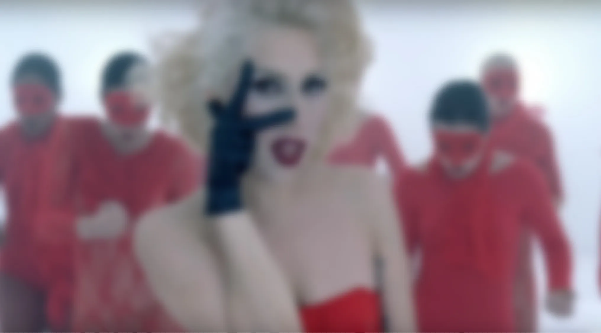 A blurry music video image of a blonde woman in a red outfit and black gloves, surrounded by people in red. Guess the music video.