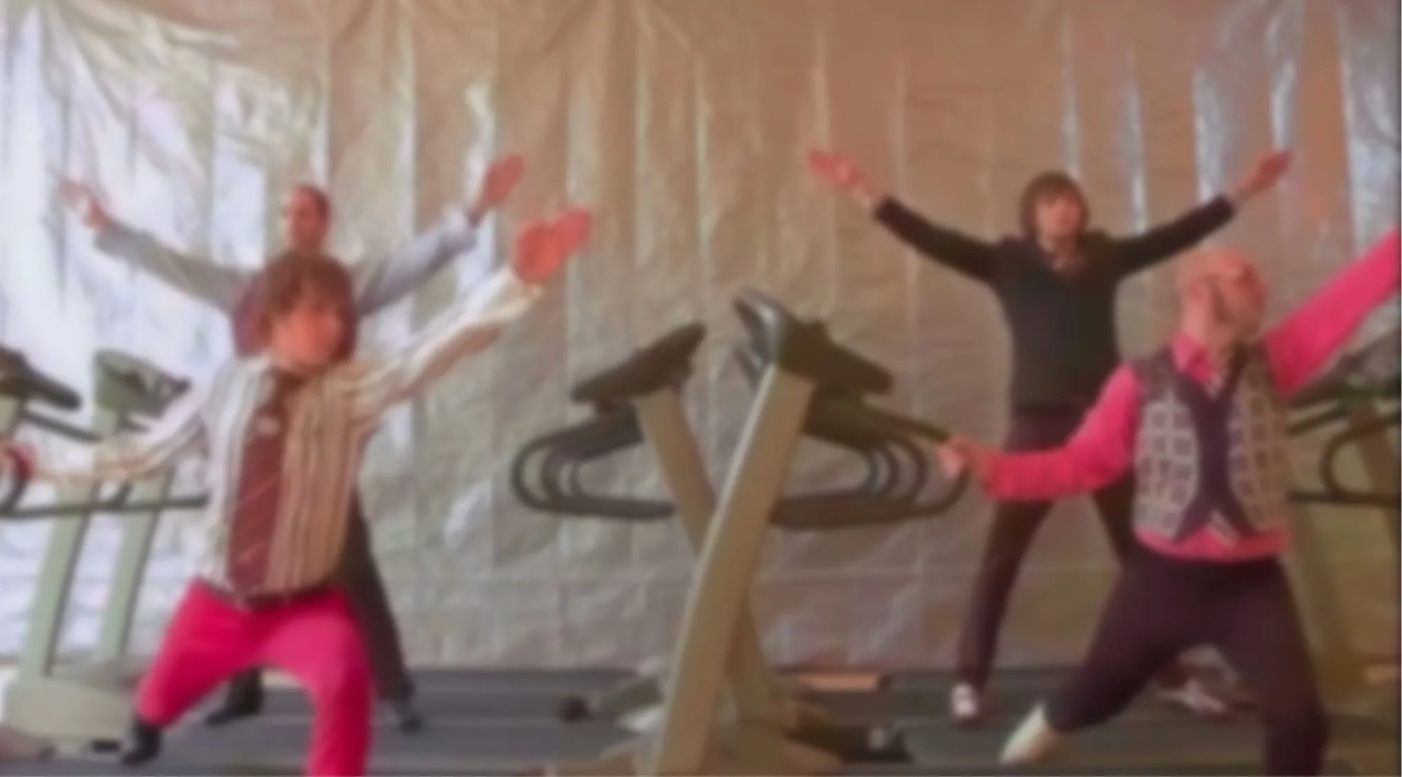 A blurry music video frame shows four people in workout clothes with arms outstretched on treadmills. Blurry music videos.