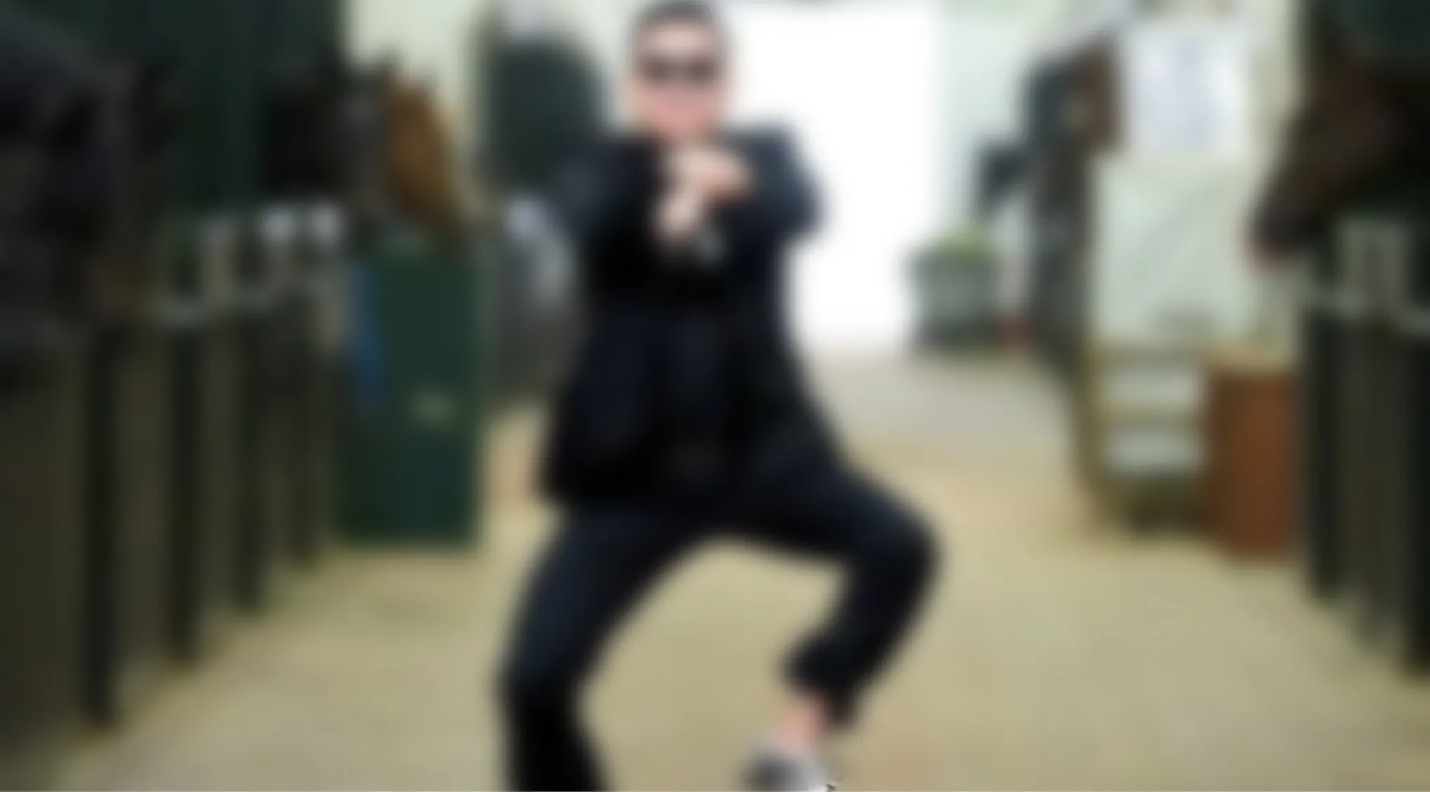 A blurry music videos quiz image of a figure in a black suit and sunglasses striking a pose in a stable.