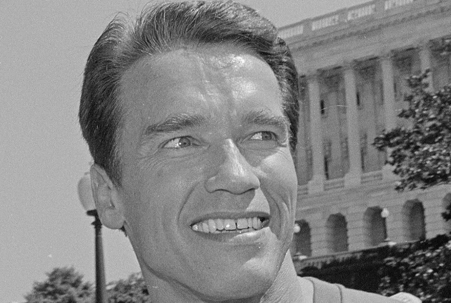 Black and white closeup of Arnold Schwarzenegger smiling with a building in the background. Bodybuilder, actor.