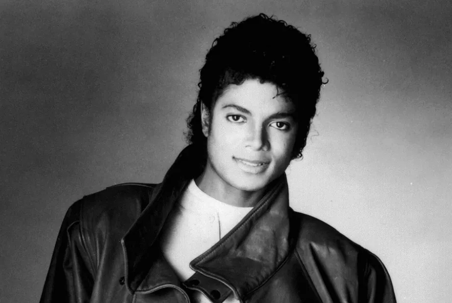 Michael Jackson, smiling and wearing a leather jacket and white shirt, captured in a black and white studio portrait.