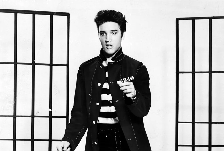 Elvis Presley in a striped prisoner shirt and dark jacket, holding a number, standing between jail cell bars.