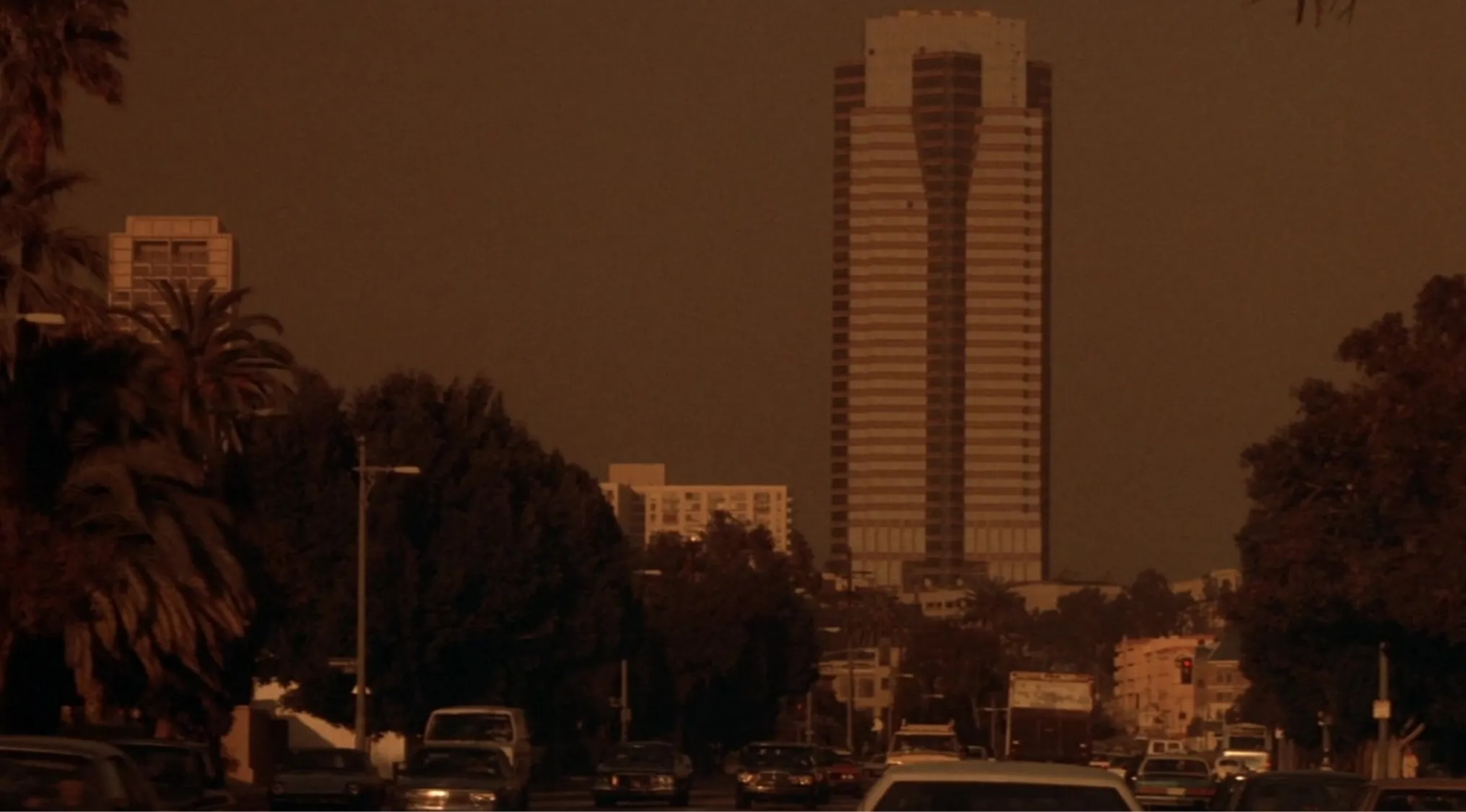 A 80s pop culture image of the Nakatomi Plaza skyscraper in Die Hard, framed by trees and cars under a hazy sky.