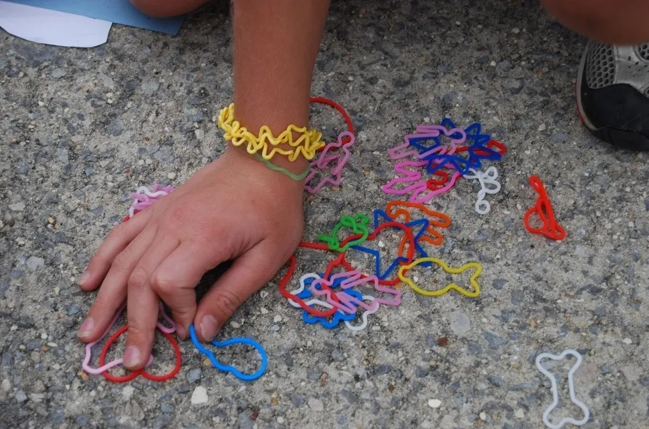 A child's hand wears neon yellow Silly Bandz while picking up other colorful, shaped elastic bands from the ground.
