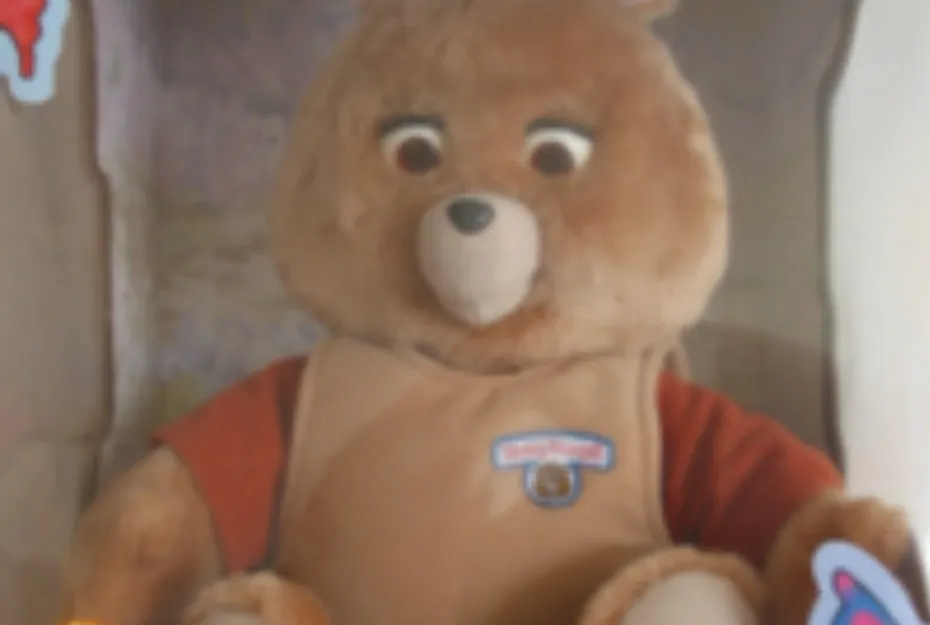 A blurry close-up of a vintage Teddy Ruxpin, a brown plush bear wearing a light brown shirt and orange undershirt.