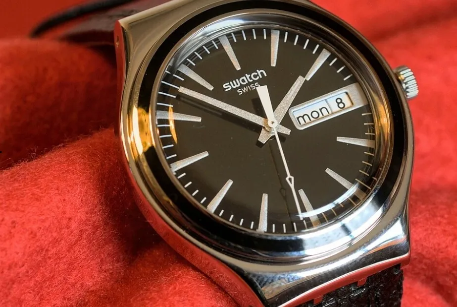 A close-up of a Swatch watch with a black face, silver hands, and a day/date display, resting on red fabric.