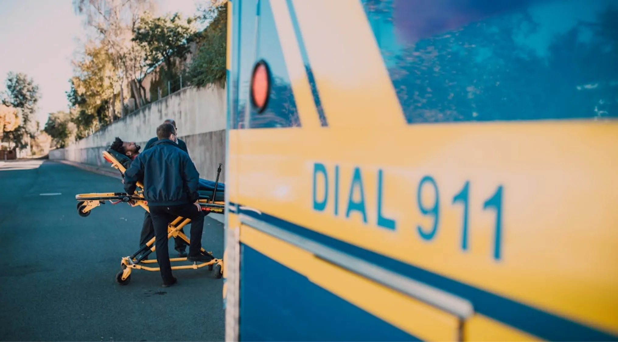 Paramedics attend to a patient on a stretcher near an ambulance with "DIAL 911" visible, emphasizing First Aid survival.
