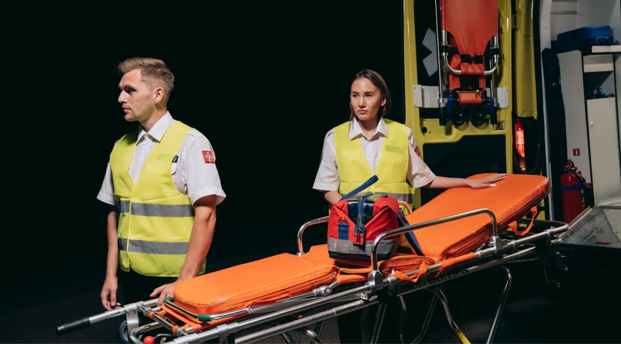 Two paramedics by an ambulance, preparing for emergency response. Essential First Aid and Survival 101 medical gear visible.