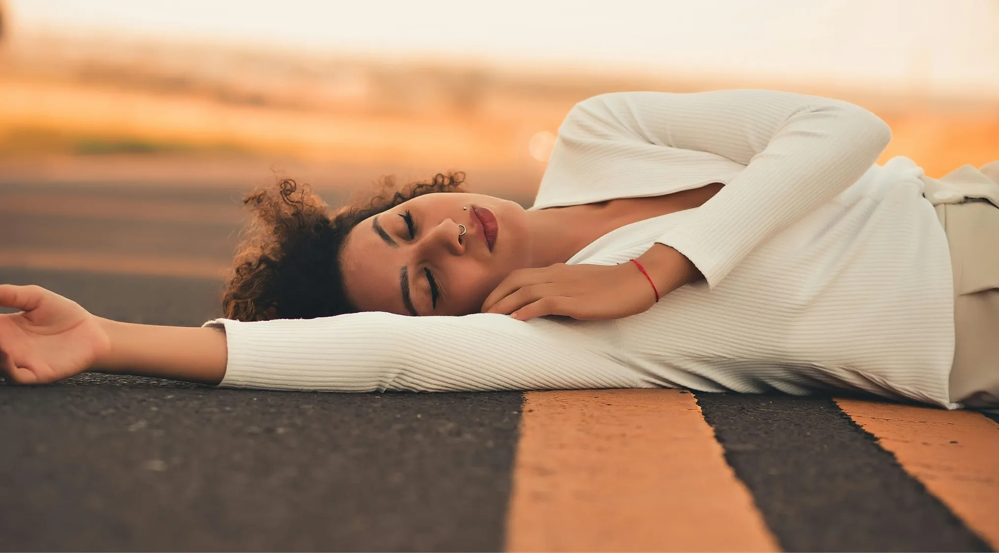 A woman with curly hair and piercings lies unconscious on an asphalt road with a yellow line, demonstrating First Aid survival 101.