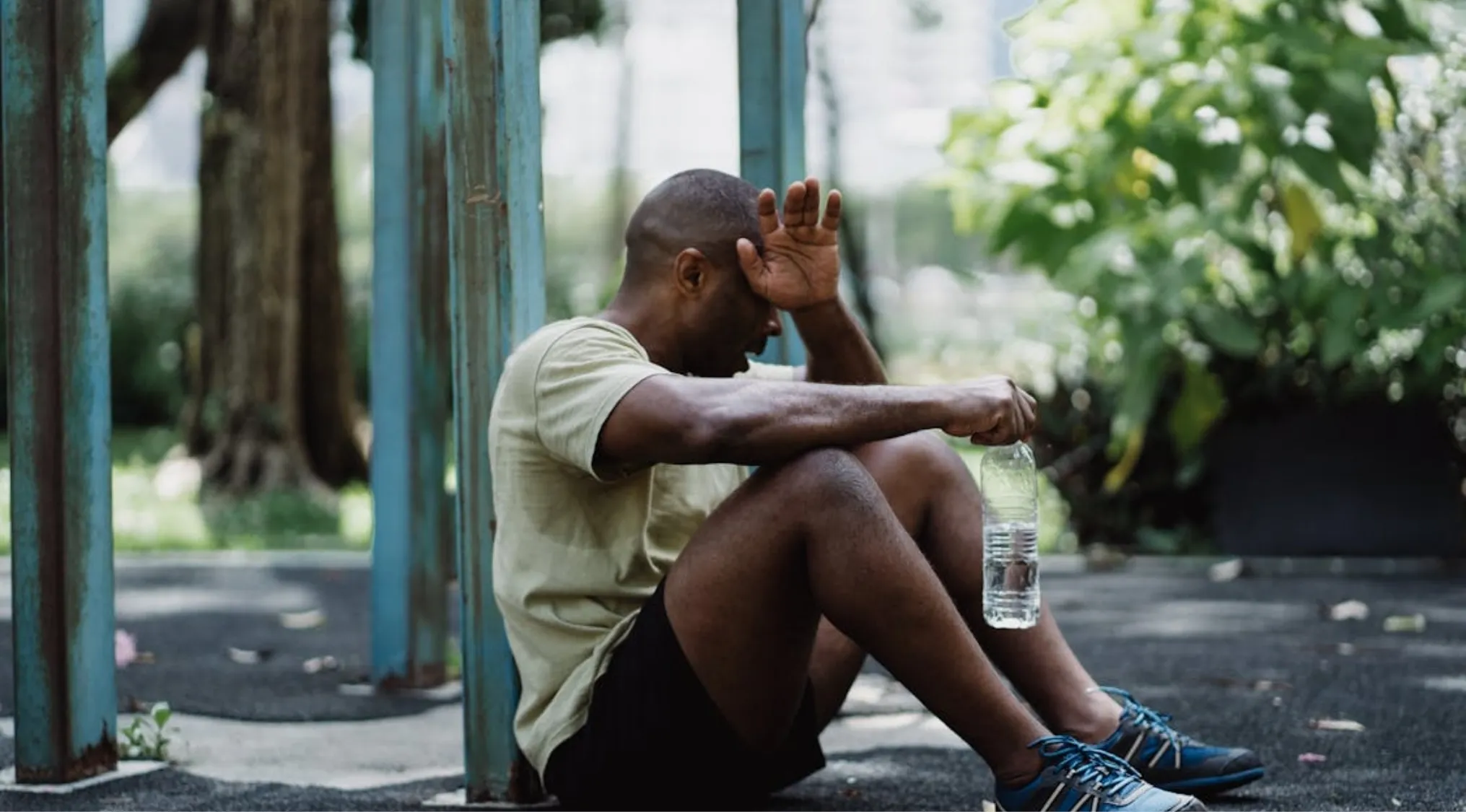 A man suffering from heatstroke rests against a blue metal structure, holding a water bottle. First Aid: Survival 101.