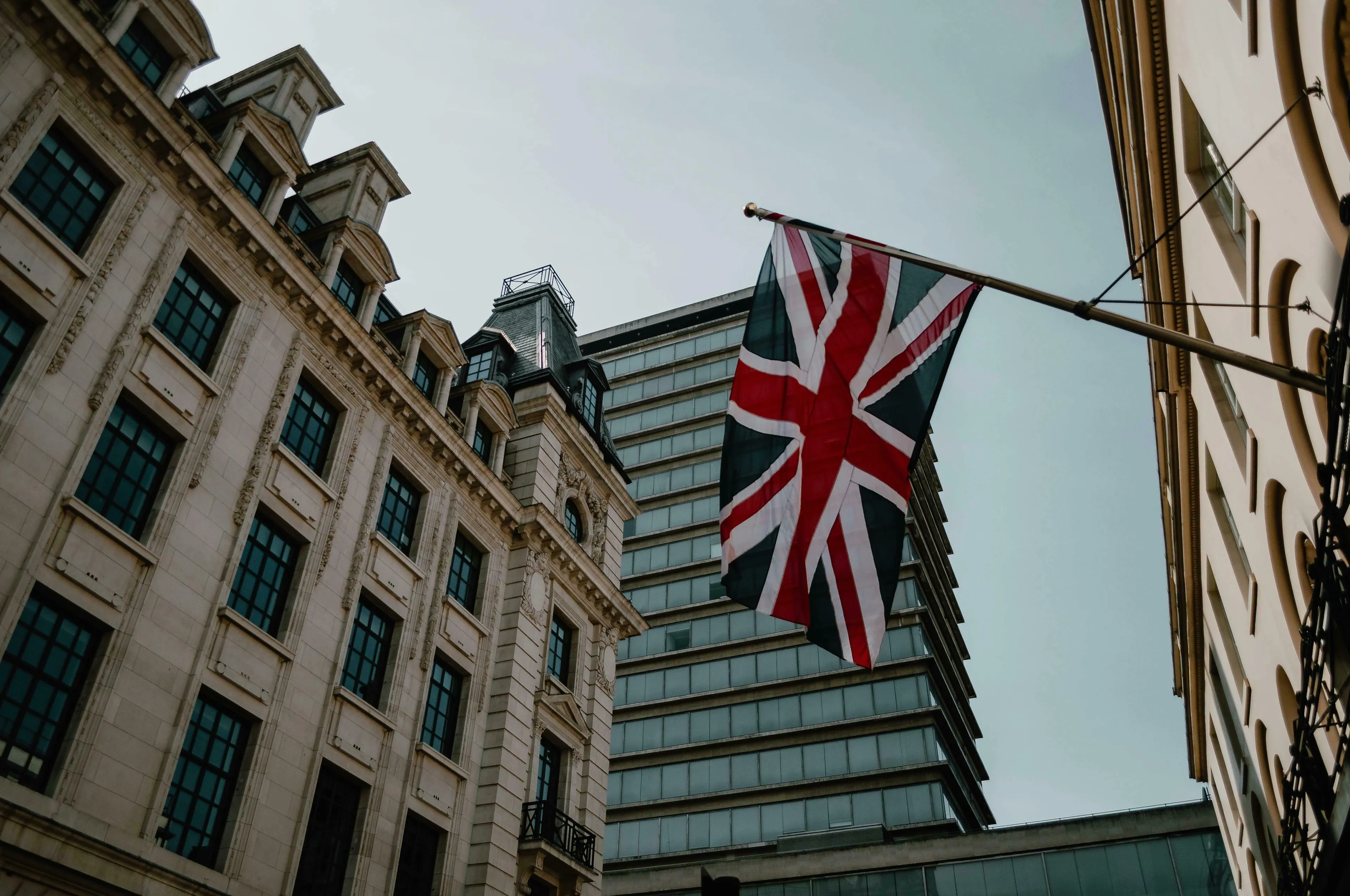A Union Jack flag hanging on a building in the UK, with other historic and modern buildings under a clear sky.