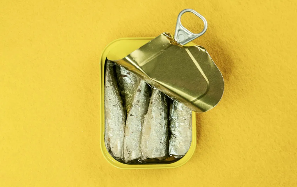 An open can of sardines with the lid pulled back, revealing small fish packed tightly in oil, on a yellow surface.