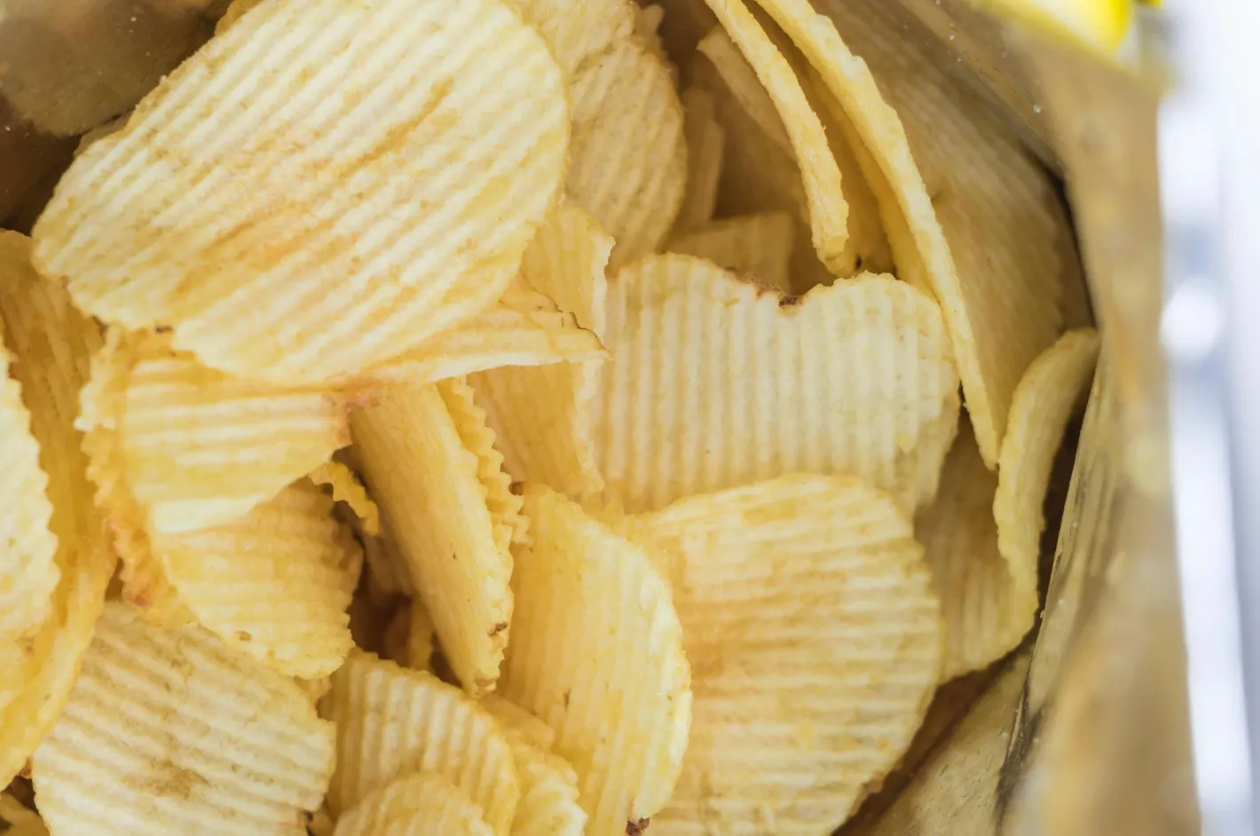 A close-up view of a packet of crisps, showing numerous golden, ridged potato chips spilling from the bag.