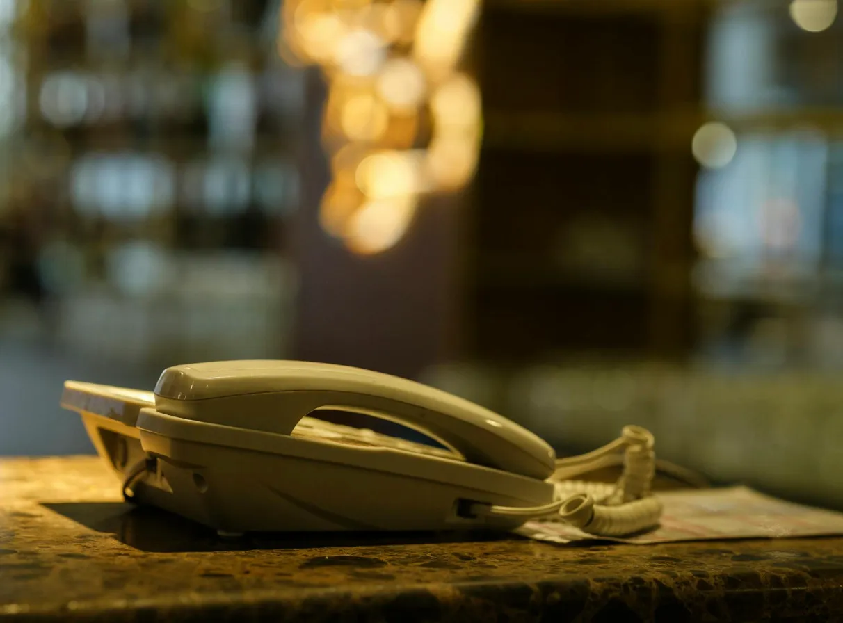 A white telephone rests on a brown counter, with a blurred background of warm lights. This is a common device like a telly.