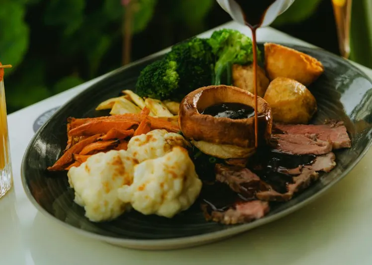 A delicious roast dinner with gravy being poured over beef, Yorkshire pudding, broccoli, cauliflower, and roasted carrots.