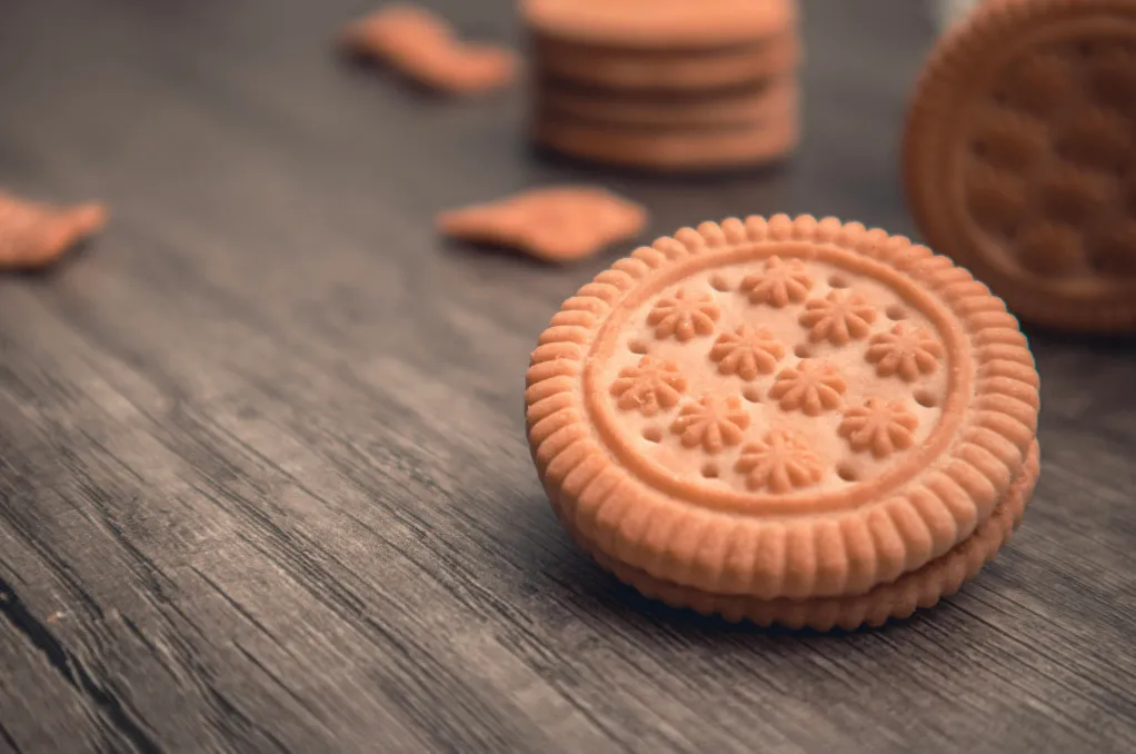 A decorative sandwich biscuit with a flower pattern on a dark wooden surface, with other biscuits blurred in the background.