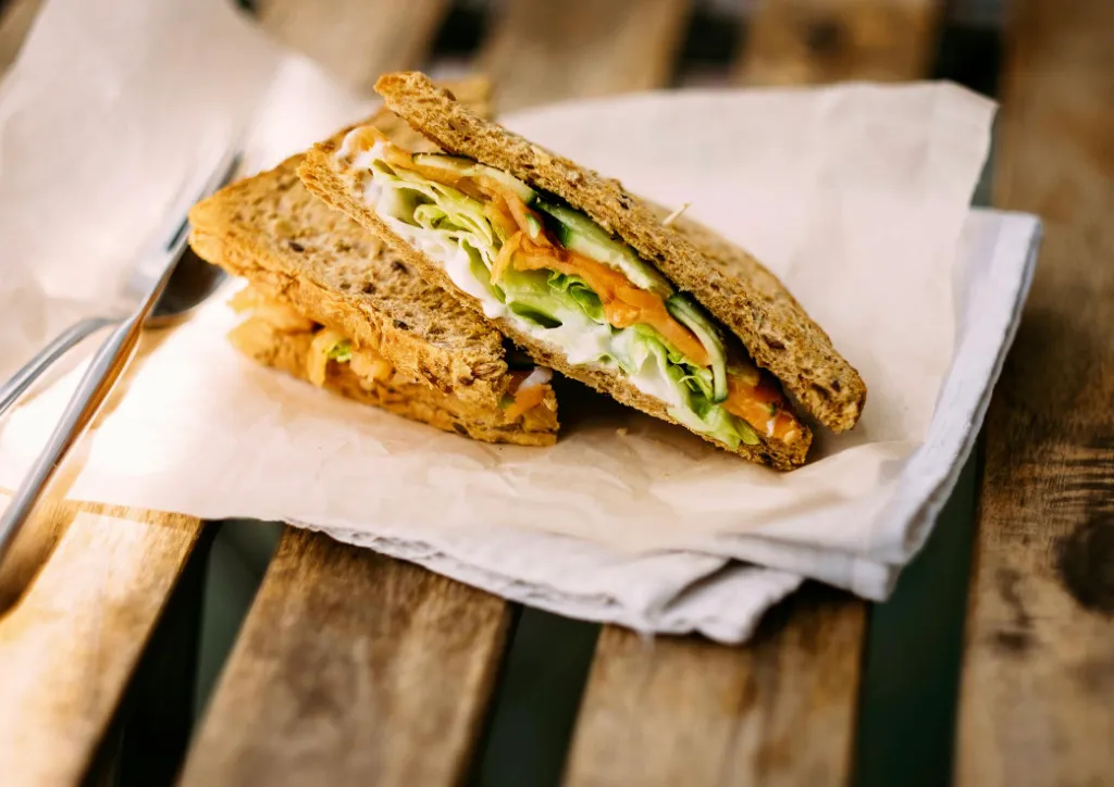 A fresh sandwich with whole wheat bread, lettuce, cucumber, and carrots, resting on parchment paper on a wooden table.