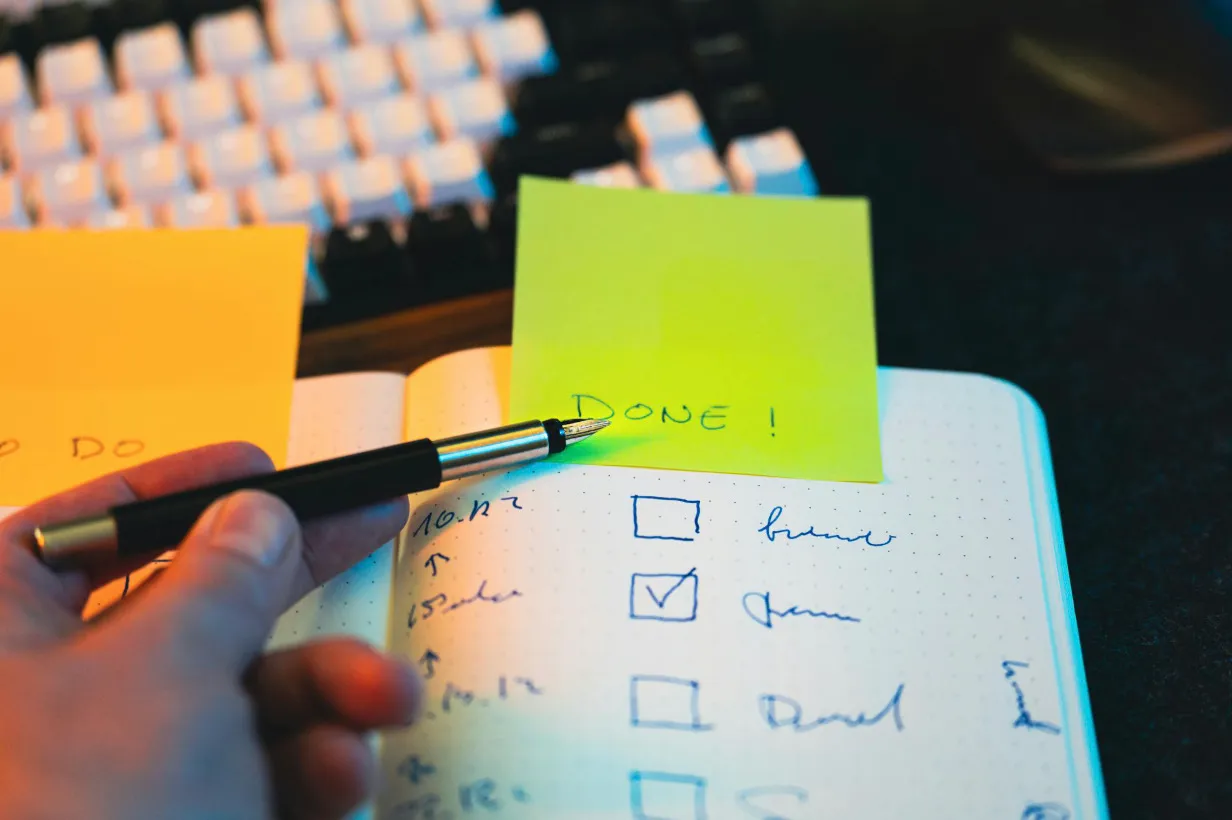 A hand holds a fountain pen, checking off tasks in a notebook with a "DONE!" sticky note. Doing small tasks with no particular goal.
