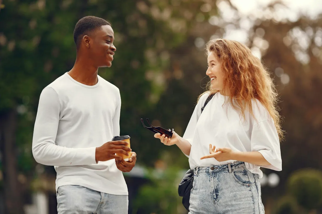 A man and woman having a friendly chat outdoors. The man holds coffee, and the woman gestures, both smiling.