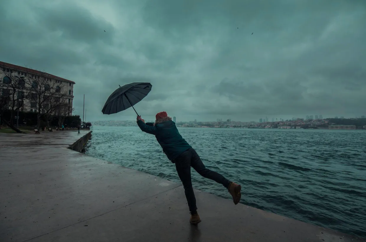 A person battling high winds with an umbrella by a stormy sea, complaining about the weather.