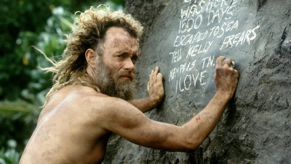Tom Hanks as Chuck Noland from Cast Away, writing messages on a rock face. He looks gaunt with a long beard and messy hair.